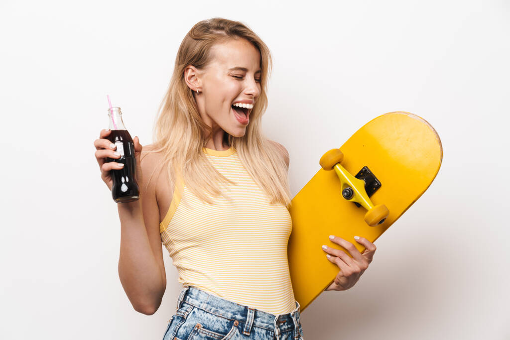 Cheery amazing young pretty woman posing isolated over white wall background holding skateboard drinking soda. - Photo, Image