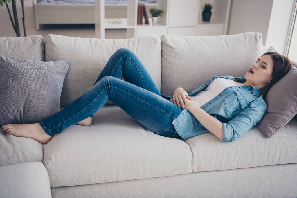 in voller Länge Foto des aufgebrachten Mädchens auf dem Sofa liegen berühren ihren Bauch leiden unter Schmerzen im Bauch haben schreckliche pms tragen Jeans-Outfit in Zimmer-Wohnung - Foto, Bild