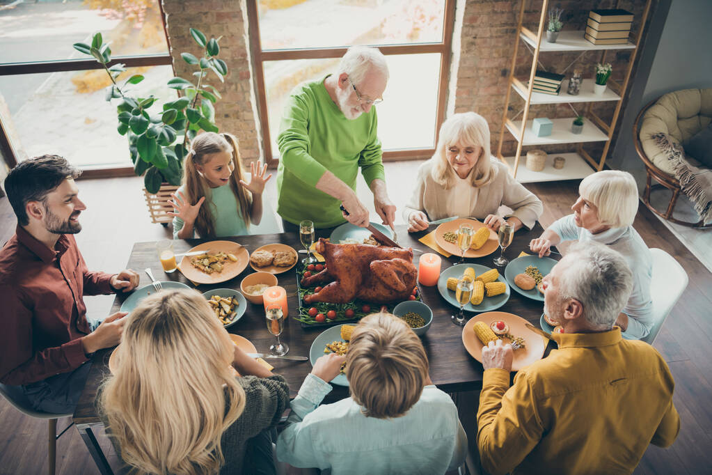 Foto de gran familia sentarse mesa de platos de banquete alrededor de pavo asado abuelo mayor haciendo rebanadas familiares hambrientos esperando emocionados en la sala de estar en el interior
 - Foto, imagen