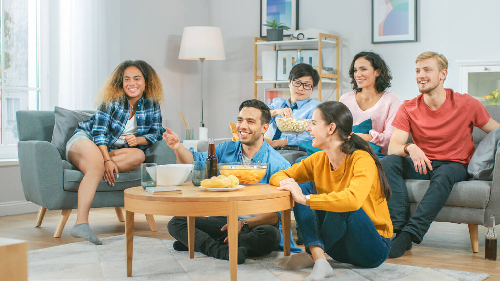 Zu Hause sitzen diverse Gruppenfreunde zusammen vor dem Fernseher, essen Snacks und trinken Getränke. Sie schauen sich wahrscheinlich Sportspiele oder Filme an. Junge Leute haben gemeinsam Spaß. - Foto, Bild