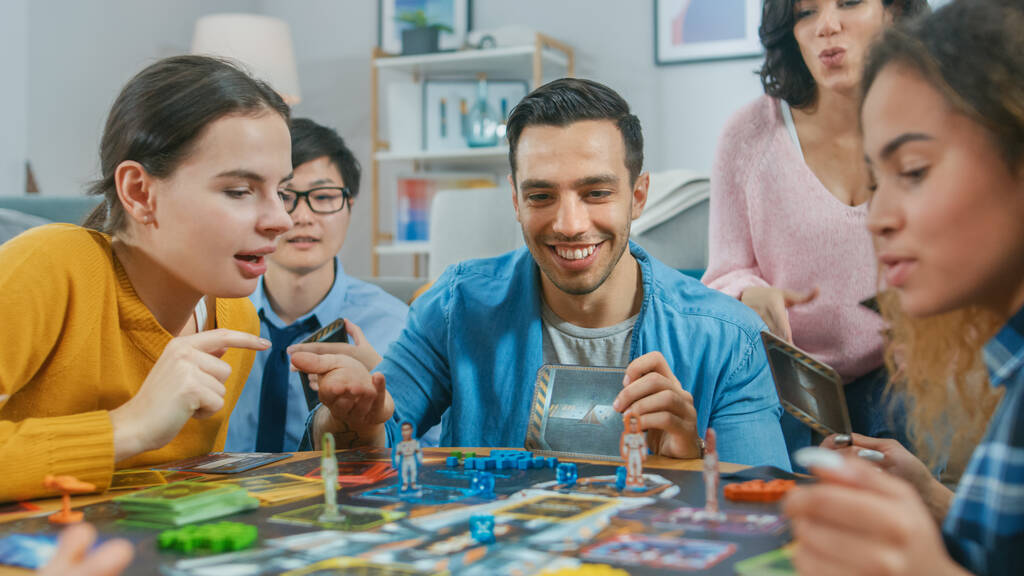 Разнообразная группа парней и девушек, играющих в стратегическую уникальную настольную игру с Cards и Dice. Друзья веселятся читая открытки, шутя, делая движения и смеясь в уютной гостиной
 - Фото, изображение