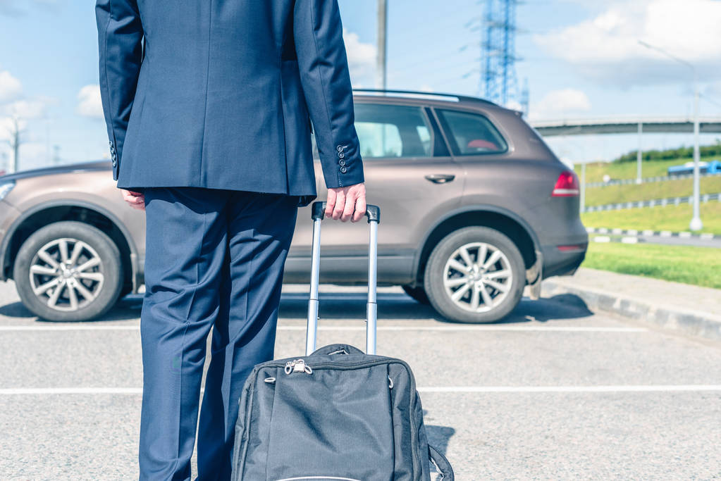 Άντρας με μπλε κοστούμι και βαλίτσα στο πάρκινγκ του αεροδρομίου στο φόντο του αυτοκινήτου του. Σχέδιο επαγγελματικού ταξιδιού - Φωτογραφία, εικόνα