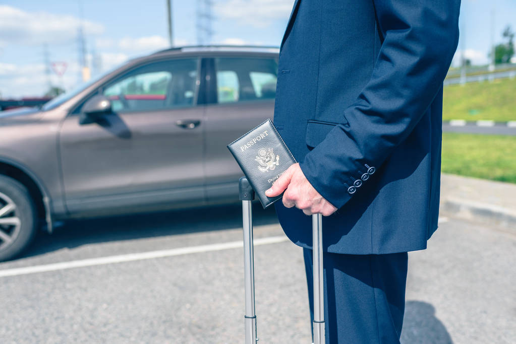 Muž v modrém obleku s kufrem a americkým pasem na letištním parkovišti na pozadí auta. Koncept služební cesty - Fotografie, Obrázek