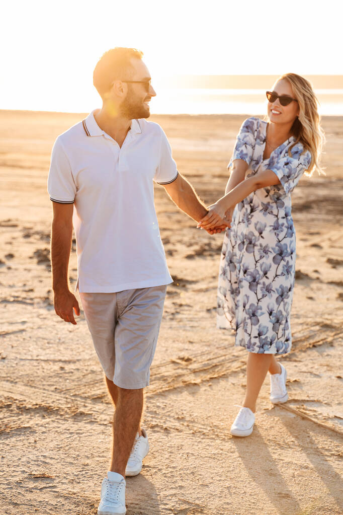 Fotografie šťastného mladého páru s úsměvem a držící se za ruce při chůzi na slunné pláži - Fotografie, Obrázek