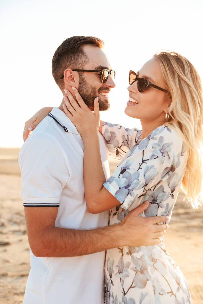 Foto de casal jovem sorridente abraçando e acariciando uns aos outros enquanto caminhava na praia ensolarada
 - Foto, Imagem