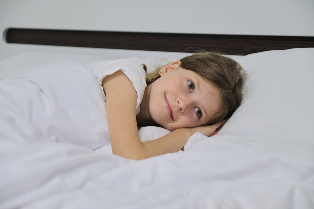 lächelndes schönes Kindermädchen auf einem Kissen liegend, weißes Bett, Gesicht aus nächster Nähe - Foto, Bild