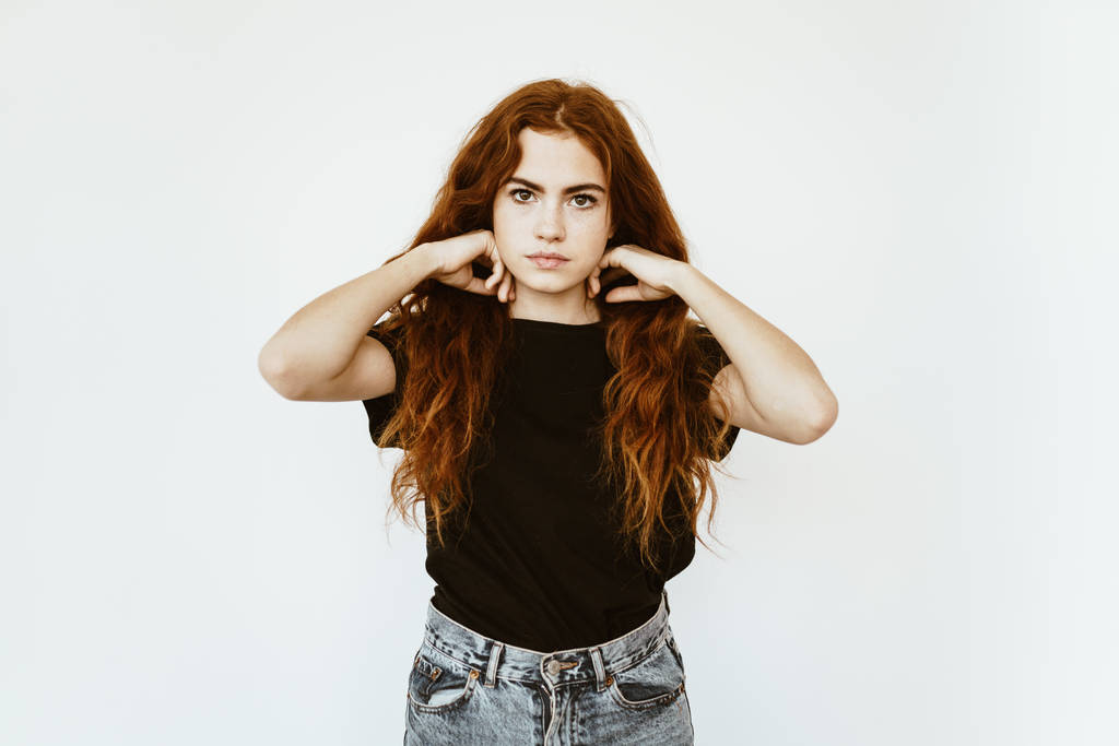 Linda joven con el pelo largo y rizado rojo con pecas en la cara en la camiseta negra y jeans en el fondo blanco endereza su cabello
 - Foto, imagen