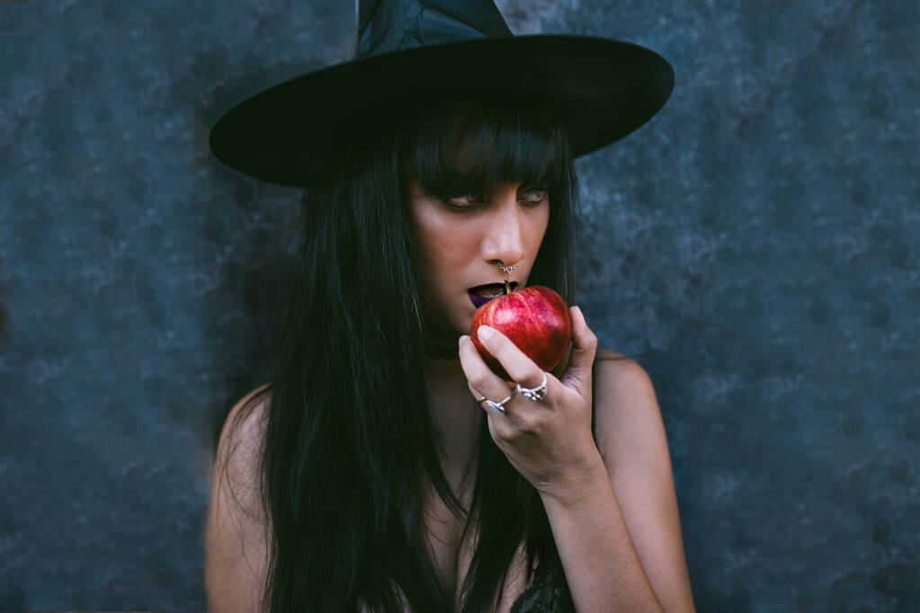 Nuori halloween-naismuotokuva syö punaista omenaa. Kauneus vihainen vampyyri noita nainen musta suu pimeydessä, yllään noita hattu. Salaperäinen Vakava malli tyttö Halloween muodostavat. Isä.
 - Valokuva, kuva