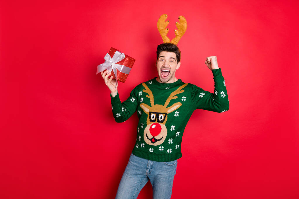 Портрет его красивый привлекательный сверх радостный веселый веселый парень в оленьем свитере, держа в руке подарок радуясь изолированы над ярким ярким блеском ярко-красного цвета фона
 - Фото, изображение
