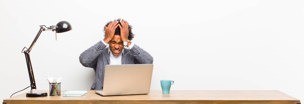 νεαρός μαύρος επιχειρηματίας που αισθάνεται άγχος και άγχος, κατάθλιψη και απογοήτευση με έναν πονοκέφαλο, σηκώνοντας και τα δύο χέρια για να το κεφάλι σε ένα γραφείο - Φωτογραφία, εικόνα
