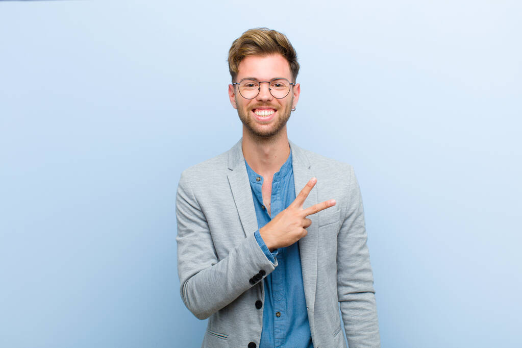 junger Geschäftsmann, der sich glücklich, positiv und erfolgreich fühlt, mit einer von Hand gefertigten V-Form über der Brust, die Sieg oder Frieden vor blauem Hintergrund zeigt - Foto, Bild