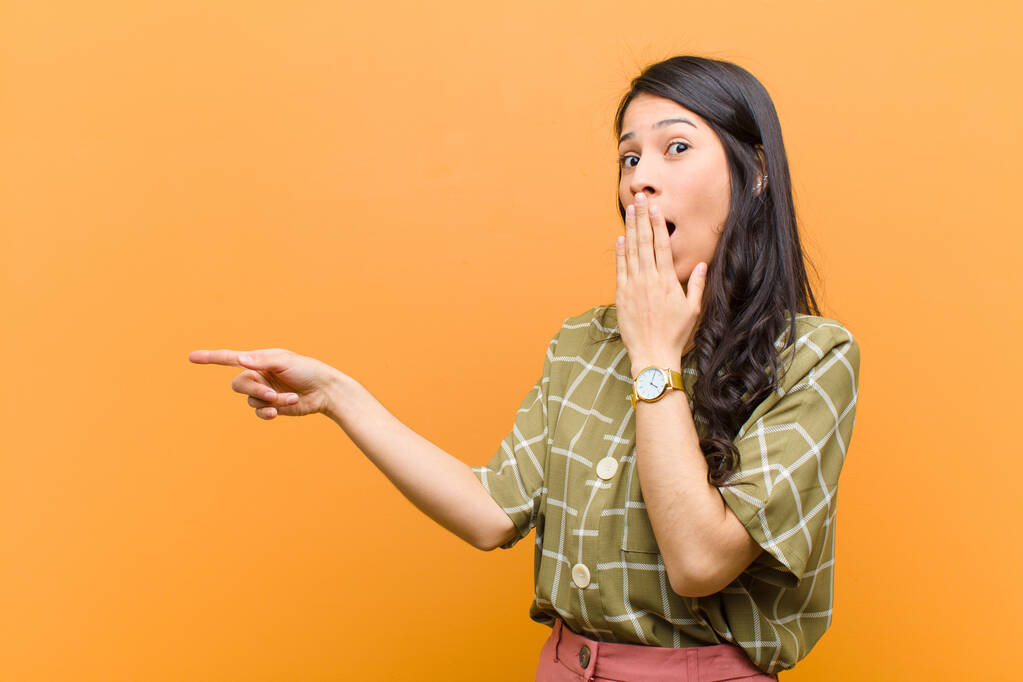 молодая довольно латиноамериканская женщина чувствует себя счастливой, шокированной и удивленной, закрывая рот рукой и указывая на боковое пространство для копирования у коричневой стены
 - Фото, изображение