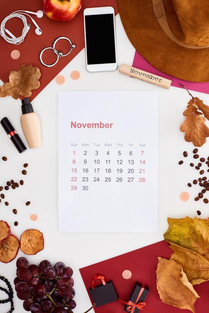 november calendar page near hat, dry leaves, fruits, donut, cosmetics, coffee grains and wooden block with november inscription isolated on white - Photo, Image