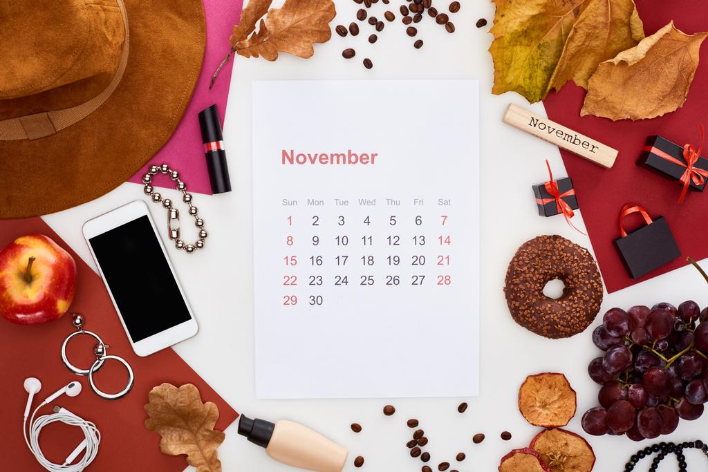 November-Kalenderseite, Hut, Smartphone, Früchte, Kosmetik, trockene Blätter, Donut, buntes Papier, Holzblock mit Novemberaufschrift isoliert auf weiß - Foto, Bild