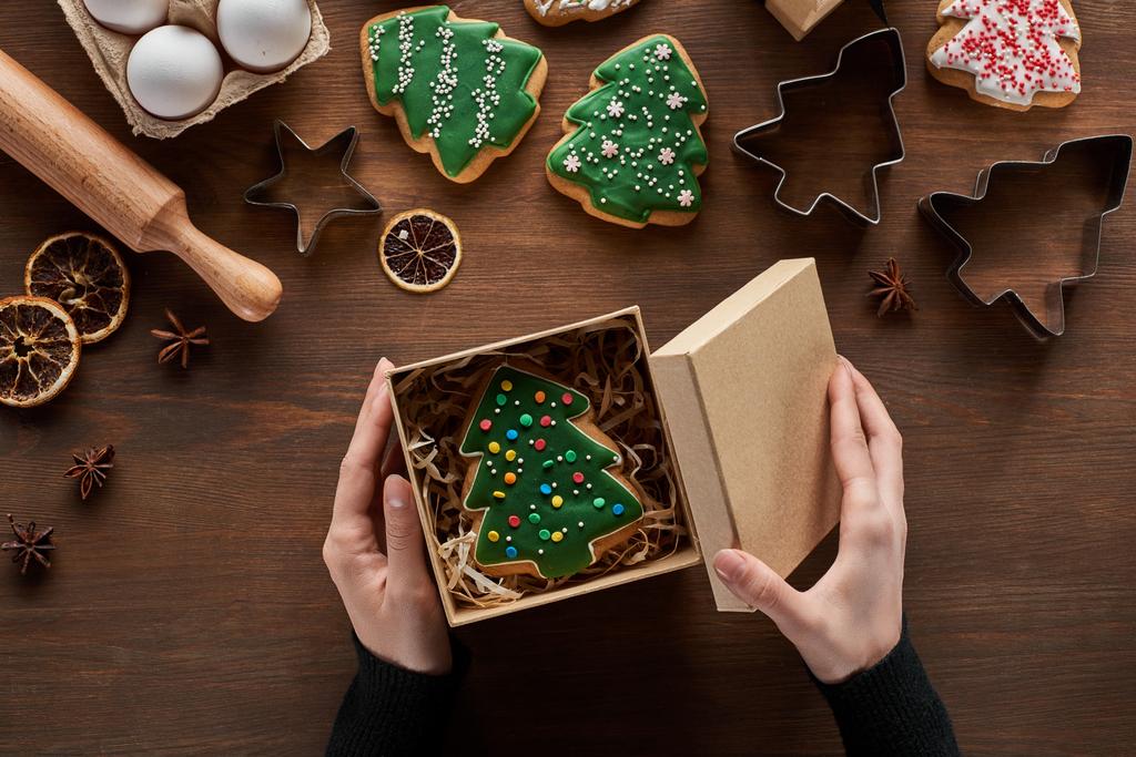 vista cortada da caixa de presente de fechamento da mulher com biscoito de árvore de Natal na mesa de madeira
 - Foto, Imagem