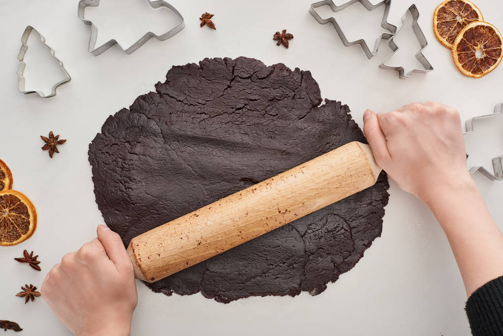 cropped view of woman holding rolling pin on raw dough for chocolate Christmas cookies on white background near anise, dough molds and dried citrus - Photo, Image