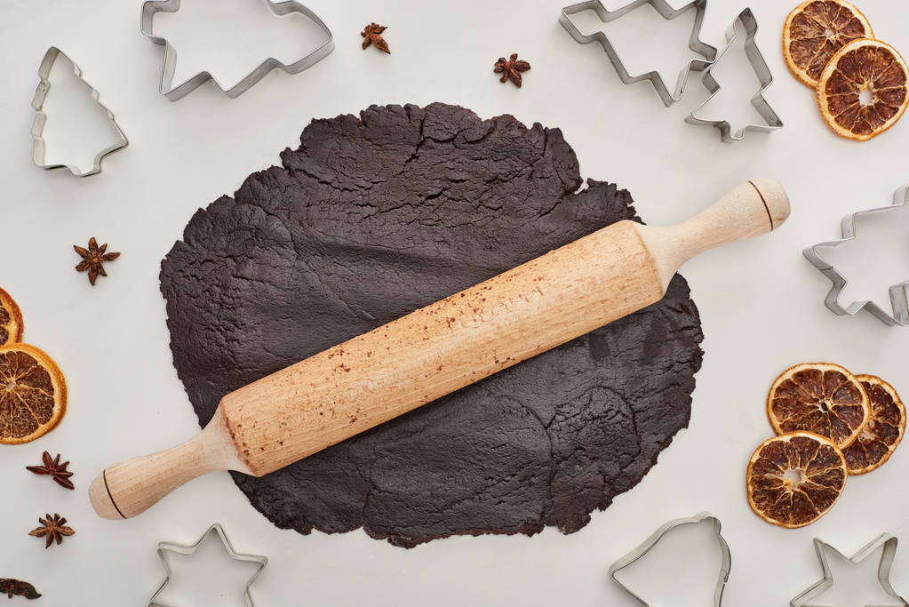 vista superior da massa crua para biscoitos de Natal de chocolate com rolo de pino no fundo branco perto de anis, moldes de massa e citrinos secos
 - Foto, Imagem