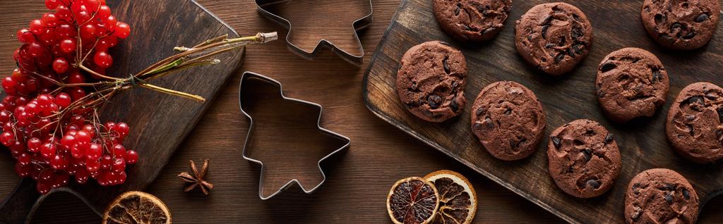 vista superior de biscoitos de chocolate perto de moldes de massa de Natal, viburnum e especiarias na mesa de madeira, tiro panorâmico
 - Foto, Imagem