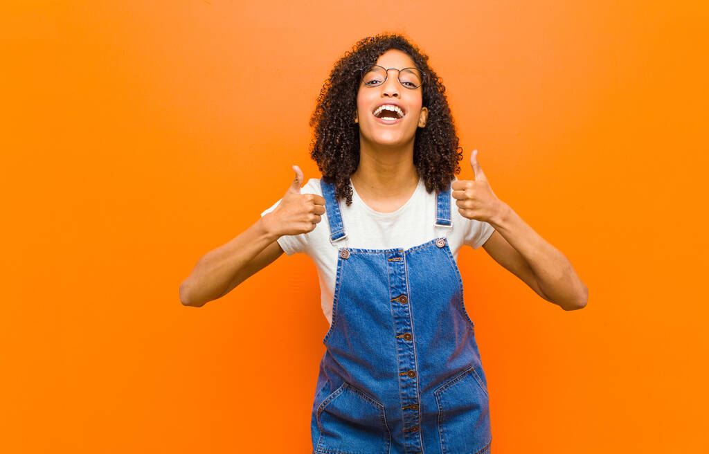 young pretty black woman smiling broadly looking happy, positive, confident and successful, with both thumbs up against orange wall - Photo, Image