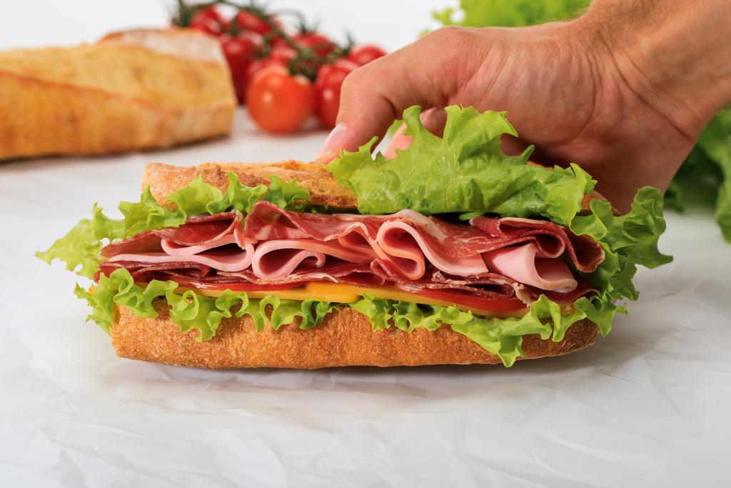 обрізаний вид людини, що тримає свіжий бутерброд з салатом, шинкою, сиром, беконом та помідорами на текстурованому білому тлі
 - Фото, зображення