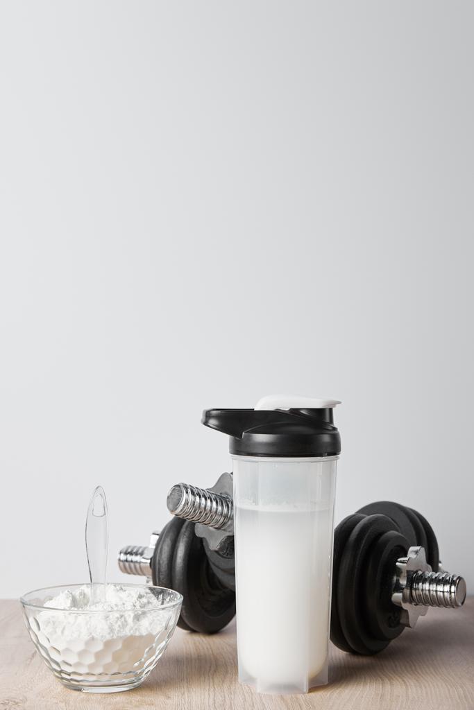 butelka sportowa z proteinowym shake 'iem w pobliżu czarnych hantli i szklanej miski z proteinowym shake' iem izolowanym na szaro - Zdjęcie, obraz