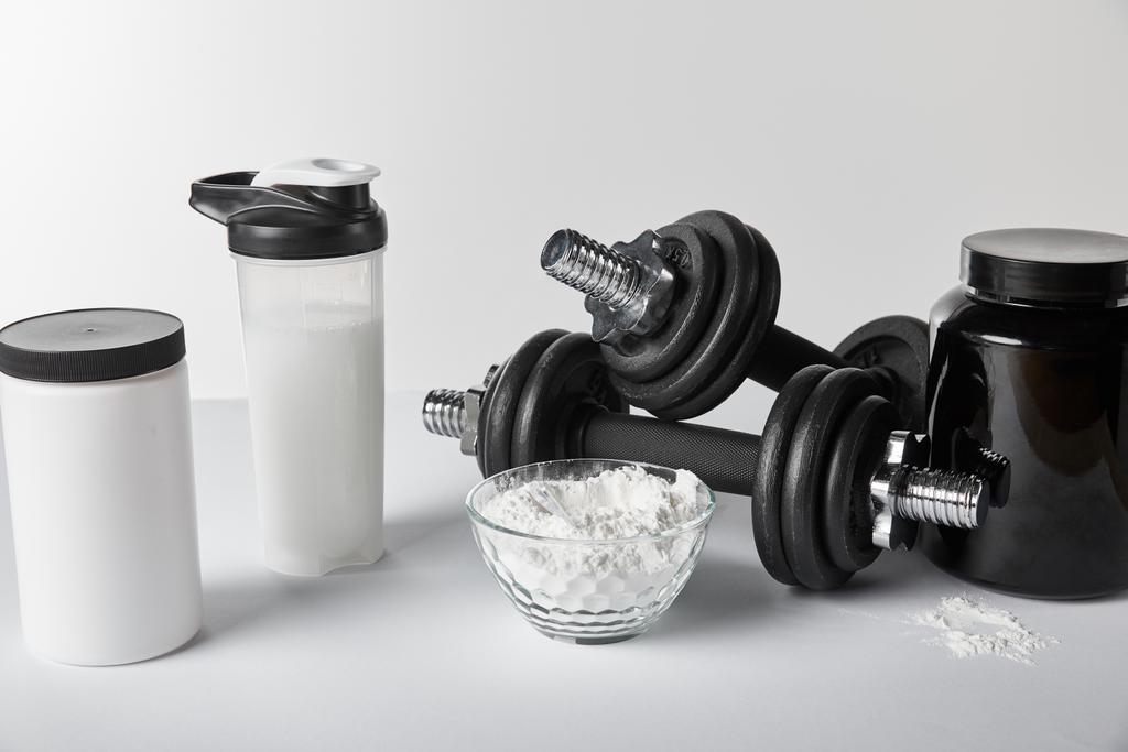 банки и спортивная бутылка возле миски с белковым порошком и гантели на белом
  - Фото, изображение