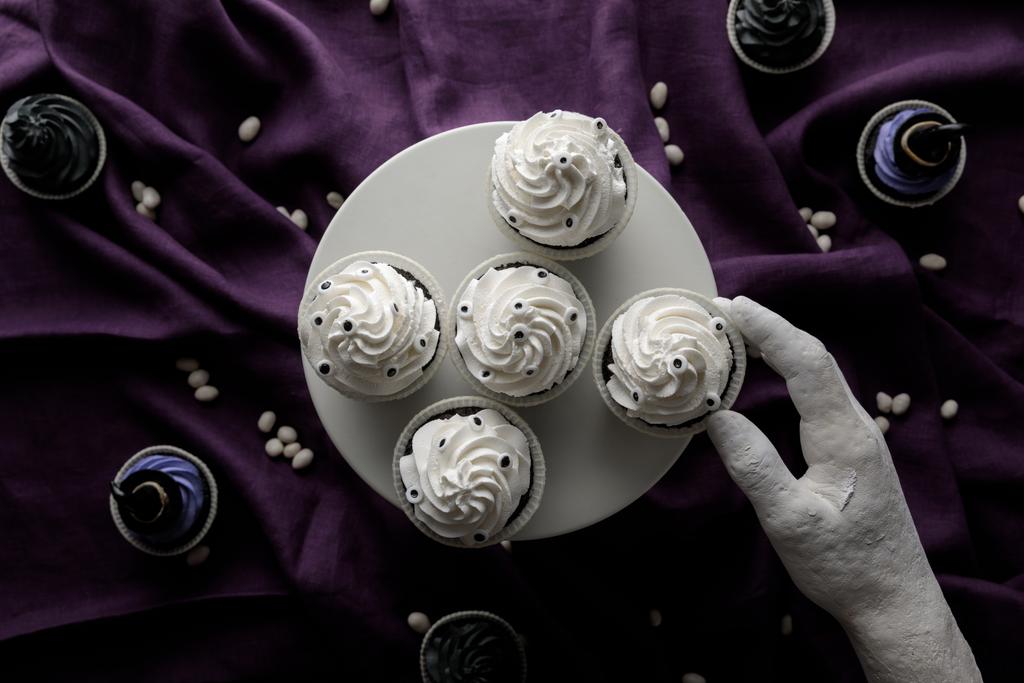 bovenaanzicht van decoratieve hand in de buurt staan met heerlijke Halloween cupcakes op paarse doek - Foto, afbeelding