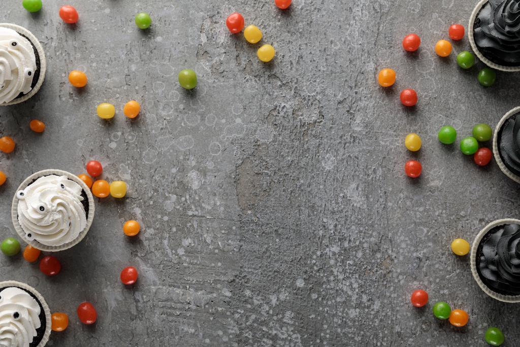 Draufsicht auf köstliche Halloween-Cupcakes mit verstreuten bunten Bonbons auf betongrauer Oberfläche - Foto, Bild