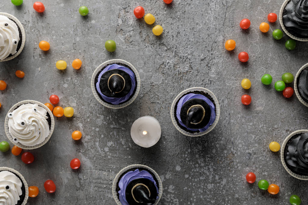 bovenaanzicht van heerlijke Halloween cupcakes en brandende kaars met verspreide kleurrijke bonbons op betonnen grijs oppervlak - Foto, afbeelding