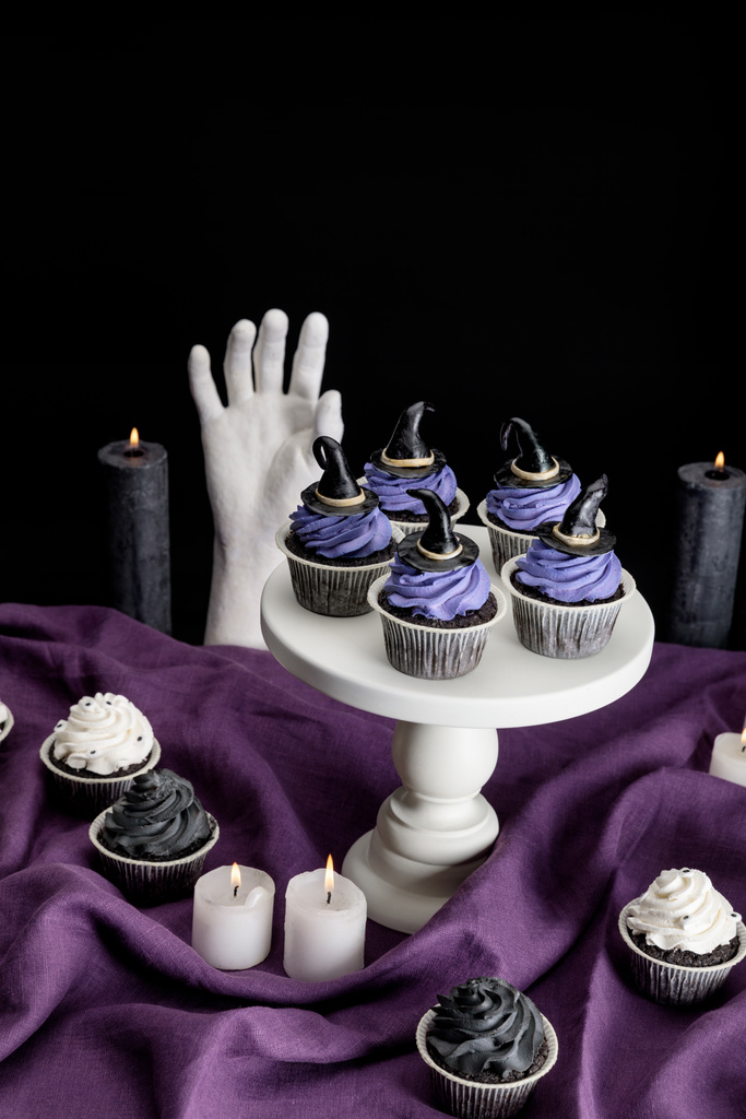 smakelijke Halloween cupcakes op witte standaard in de buurt van brandende kaarsen en decoratieve hand op paarse doek geïsoleerd op zwart - Foto, afbeelding