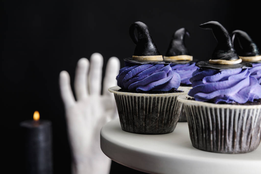 selectieve focus van smakelijke Halloween cupcakes met blauwe room en decoratieve heksenhoeden op witte standaard geïsoleerd op zwart - Foto, afbeelding