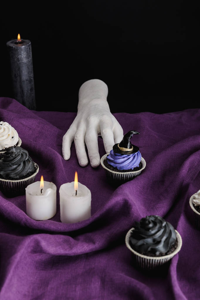 Dekoratif el, cadılar bayramı keklerinin yanında ve mor kumaşın üzerinde yanan mumlar, siyah üzerine izole edilmiş. - Fotoğraf, Görsel