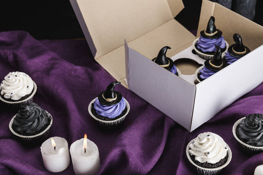 saborosos cupcakes de Halloween com creme azul e chapéus de bruxa decorativos na caixa perto de velas ardentes em pano roxo
 - Foto, Imagem