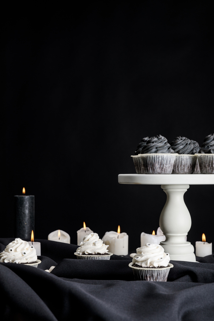 chutné Halloween cupcakes s černou smetanou na bílém stojanu v blízkosti hořící svíčky izolované na černé - Fotografie, Obrázek