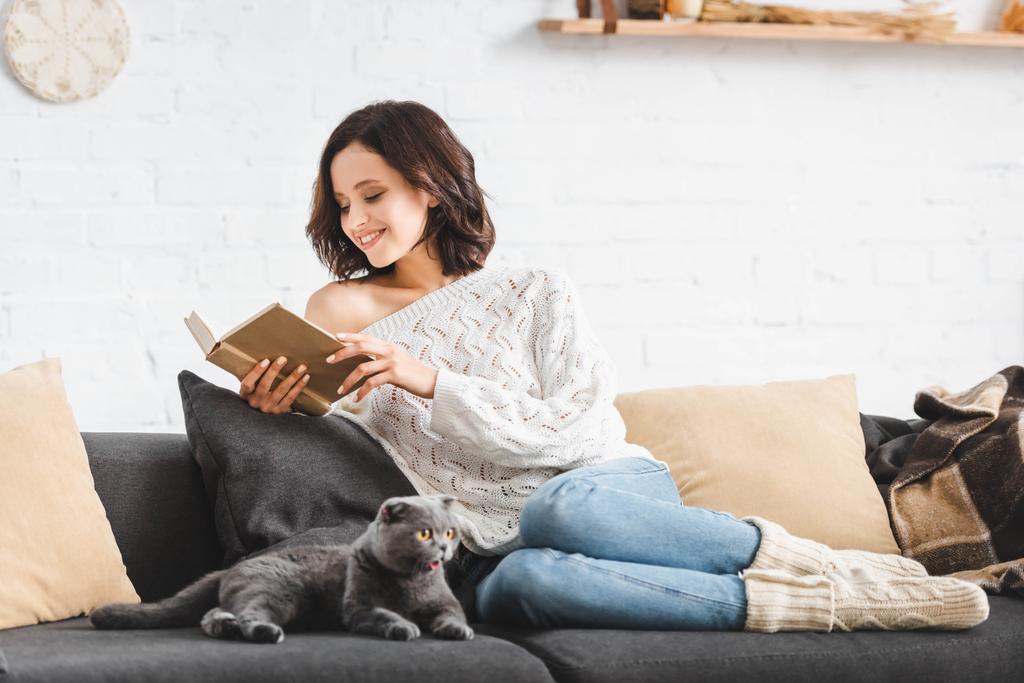 χαρούμενη γυναίκα διαβάζοντας βιβλίο με σκωτσέζικη πτυσσόμενη γάτα στον καναπέ - Φωτογραφία, εικόνα