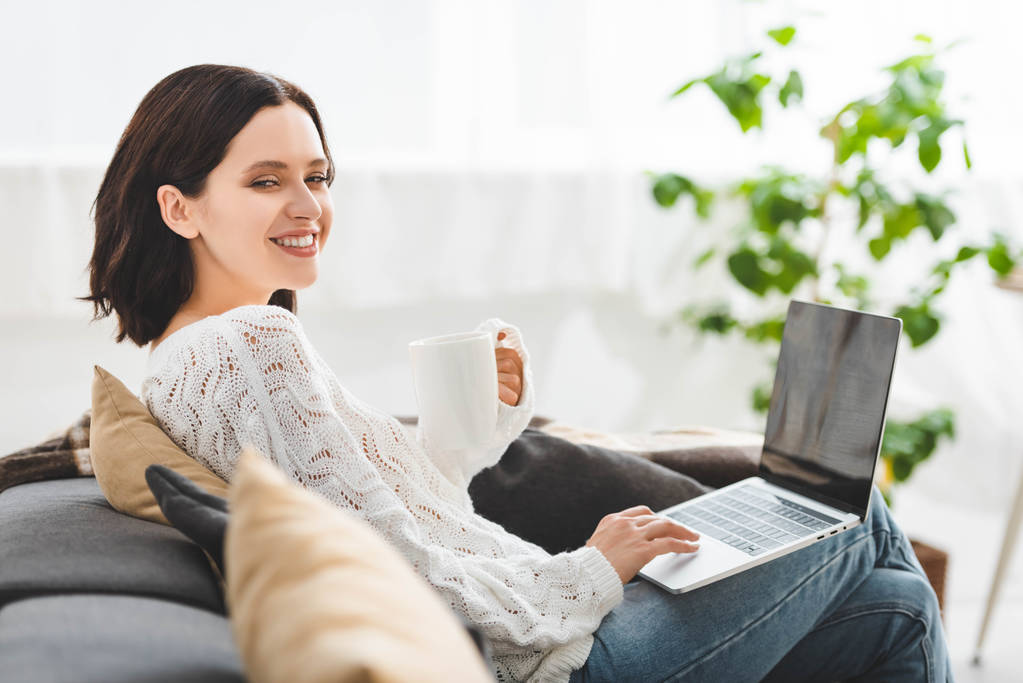 cheerful girl with cup of tea using laptop at cozy home - 写真・画像