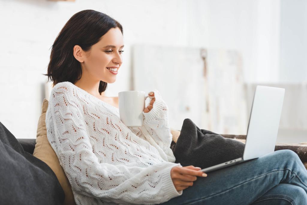 atrakcyjna uśmiechnięta dziewczyna z filiżanką kawy oglądając film na laptopie w przytulnym domu - Zdjęcie, obraz