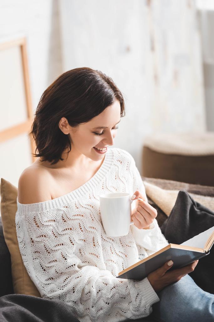 весела брюнетка дівчина читає книгу на дивані з чашкою кави
 - Фото, зображення