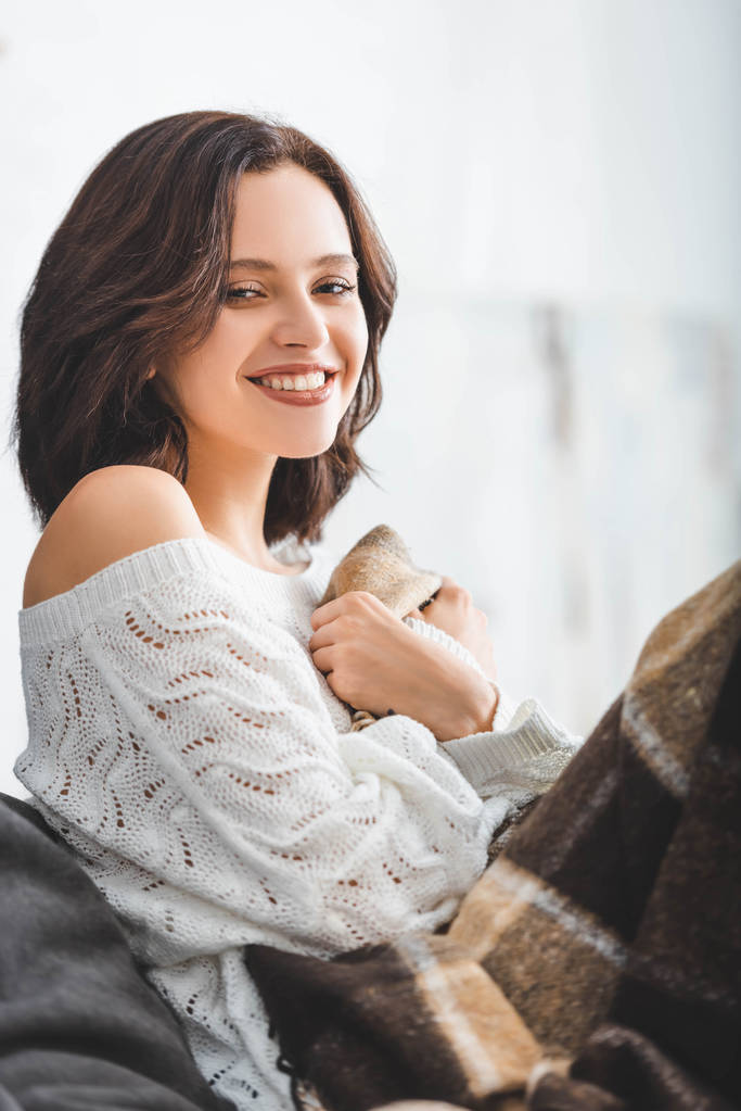 улыбающаяся брюнетка с одеялом, сидящая на диване в уютной гостиной
  - Фото, изображение