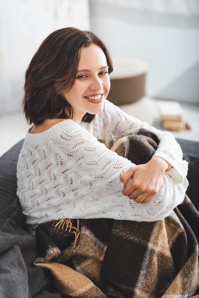menina sorridente em cobertor sentado no sofá na acolhedora sala de estar
  - Foto, Imagem