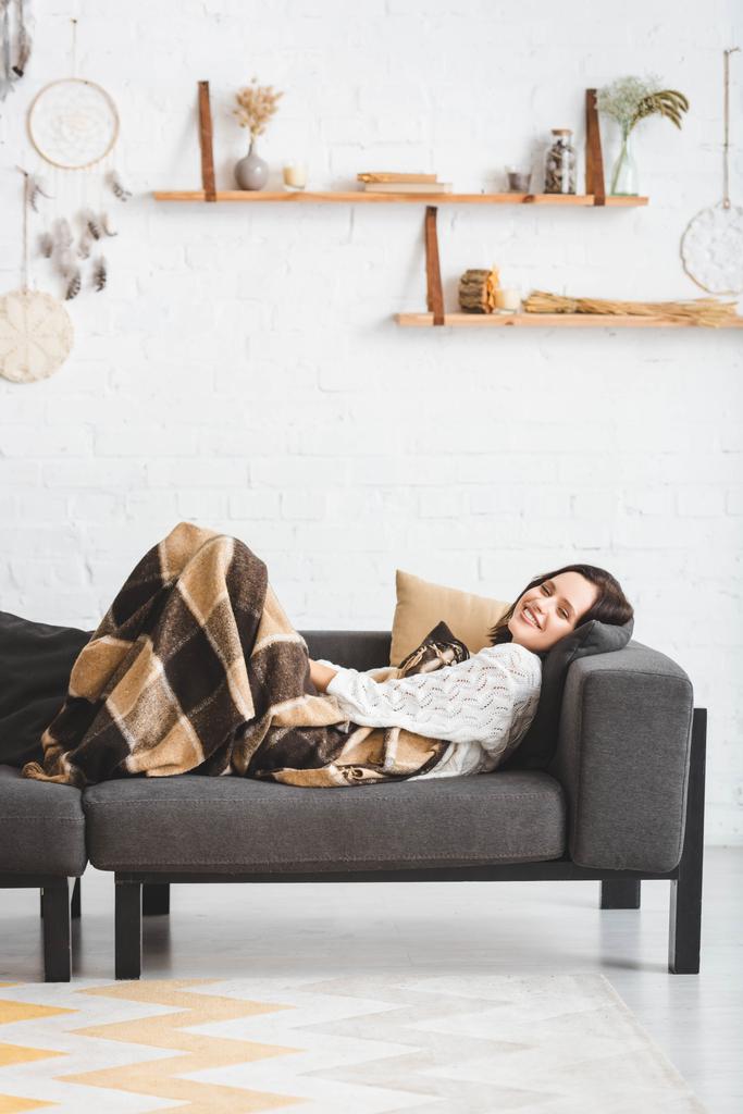 bella ragazza sorridente in coperta sdraiata sul divano in accogliente soggiorno
  - Foto, immagini