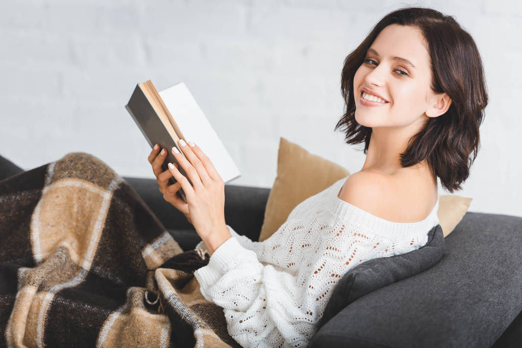 brunette glimlachende vrouw in deken lezen boek op de bank thuis - Foto, afbeelding