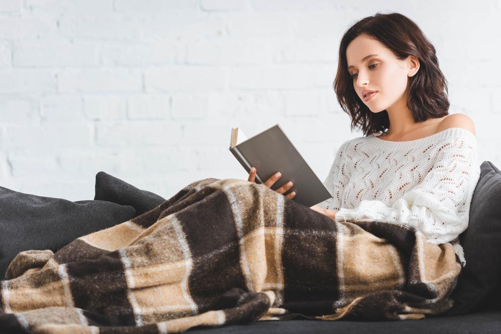 mooie jonge vrouw in deken lezen boek op de bank thuis - Foto, afbeelding