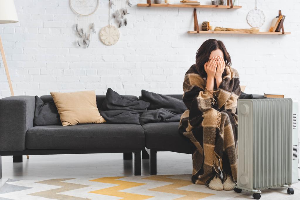 κρύο κορίτσι καλύπτει το πρόσωπό της, ενώ προθέρμανση με κουβέρτα και θερμάστρα στο σαλόνι - Φωτογραφία, εικόνα