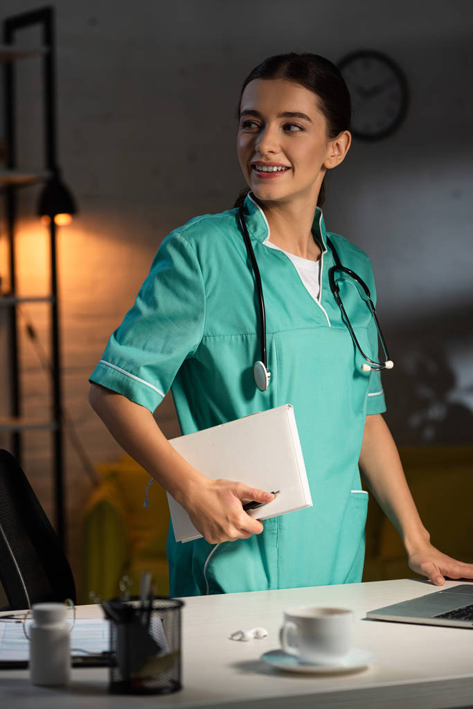 enfermera atractiva en uniforme sonriendo y sosteniendo el cuaderno durante el turno de noche
 - Foto, imagen