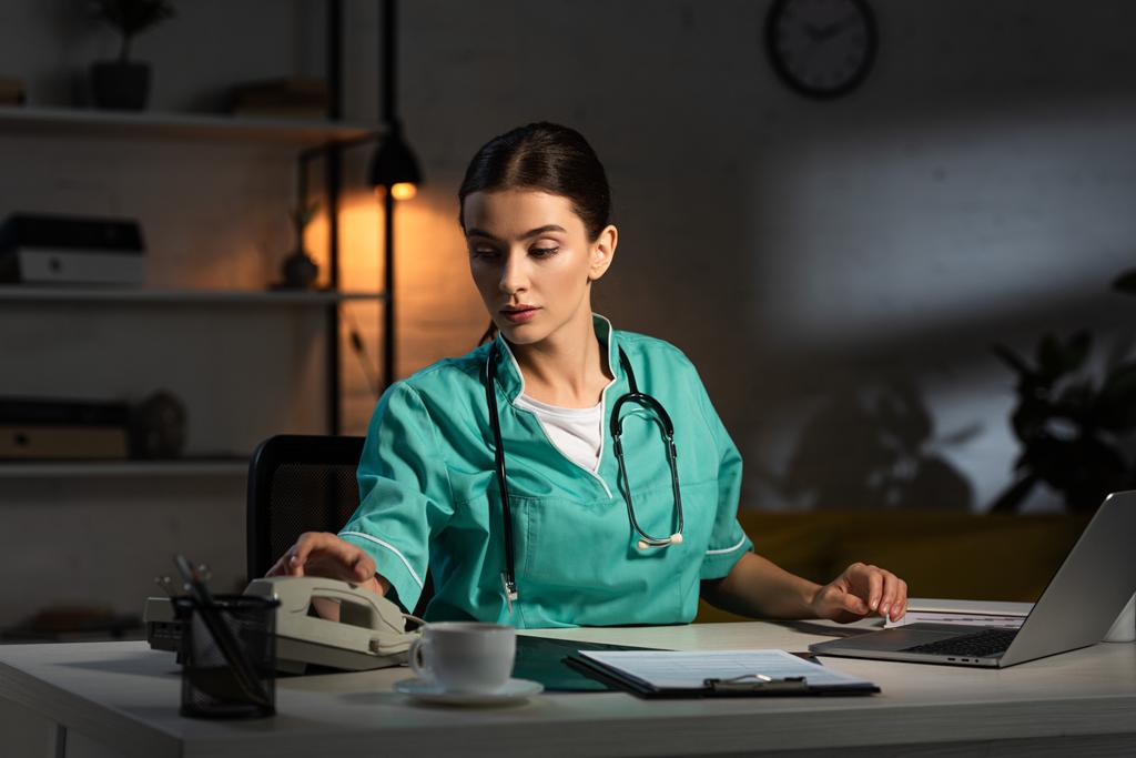 aantrekkelijke verpleegster in uniform zitten aan tafel en het nemen van handset tijdens de nachtdienst - Foto, afbeelding