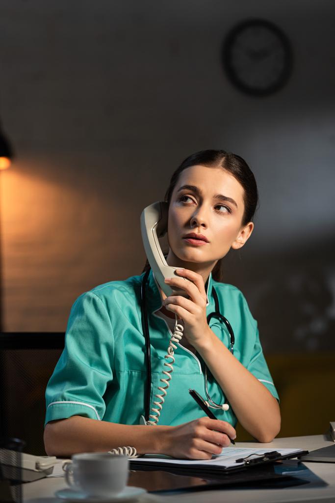 attractive nurse in uniform sitting at table and talking on telephone during night shift - Photo, Image