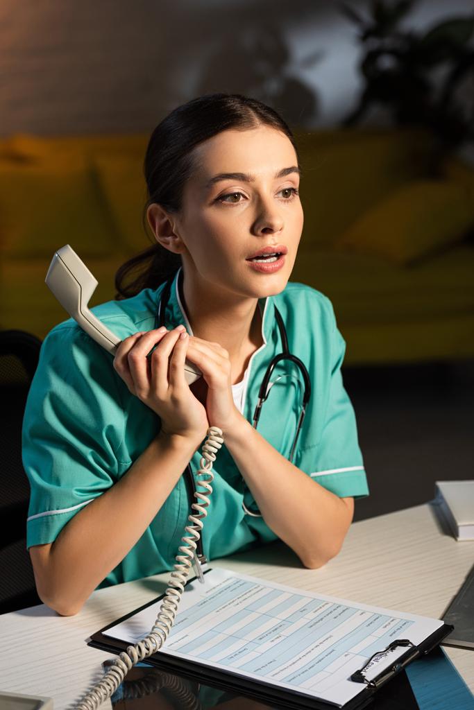 夜勤時にテーブルに座って携帯電話を持っている魅力的な看護師 - 写真・画像