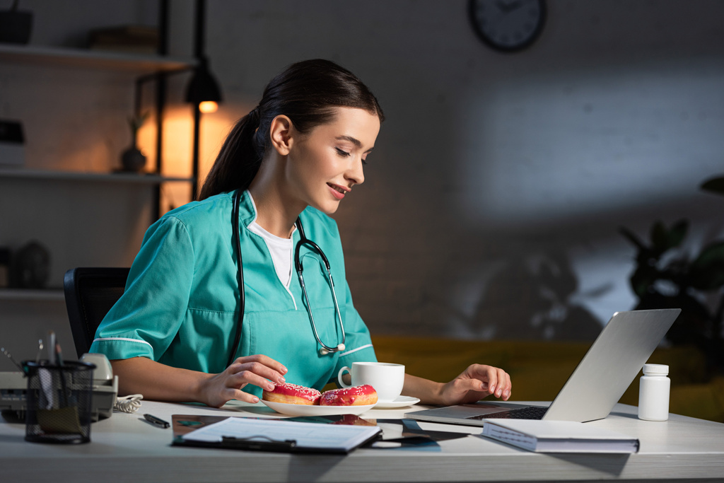 привлекательная и улыбающаяся медсестра в форме сидит за столом и смотрит на ноутбук в ночную смену
 - Фото, изображение