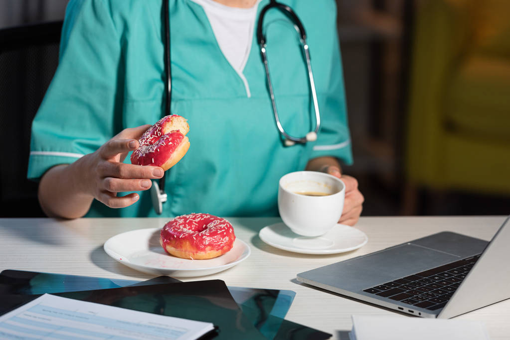 Ausgeschnittene Ansicht einer Krankenschwester in Uniform mit Donut und Tasse während der Nachtschicht - Foto, Bild